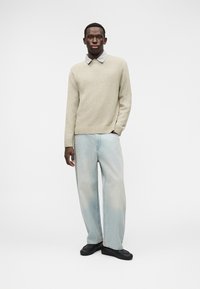 Ανοιχτό γκρι πουλόβερ πλεκτό με στρογγυλή λαιμόκοψη, φορεμένο πάνω από ένα πουκάμισο με καρό γιακά, σε συνδυασμό με φαρδιά, ανοιχτό μπλε τζιν παντελόνια και μαύρα παπούτσια.