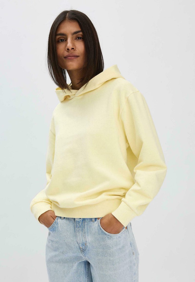 Gelbes Kapuzensweatshirt aus Baumwollmischung, mit langen Ärmeln und lockerer Passform, kombiniert mit hellblauen Jeans.