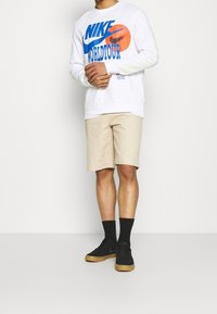 Valkoinen pitkähihainen Nike-paita, jossa on sininen ja oranssi printti, beige shortsit, mustat sukat ja mustat kengät, joissa on kumipohjat. Yksinkertainen muotoilu, rento istuvuus.