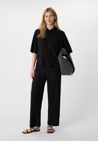 Schwarzes, zerknittertes Hemd mit kurzen Ärmeln und Brusttasche, kombiniert mit passenden weiten Hosen. Abgerundet mit einer schwarz-weißen gestreiften Tasche.