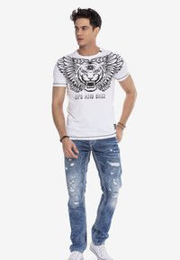 Weißes T-Shirt mit kurzen Ärmeln, das ein schwarzes Tiger-Graphic und Flügelmotiv zeigt, kombiniert mit ausgewaschenen blauen Jeans und weißen Sneakers.
