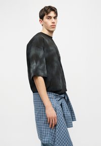 Νέος με κοντά μαλλιά που φοράει μαύρο πουκάμισο tie-dye σε μεγάλο μέγεθος και ένα μπλε καρό πουκάμισο δεμένο γύρω από τη μέση του, κοιτάζοντας πλάγια.