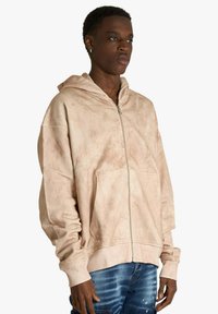 Sweat à capuche beige à fermeture éclair en tissu doux avec une finition texturée, doté d'une poche avant et d'une coupe décontractée. Porté avec un jean bleu usé.