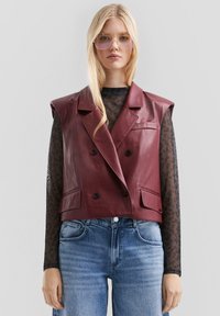 Bordeauxfarbenes Leder-Cropped-Blazer-Weste mit doppelt geknöpftem Design, spitzen Revers, zwei vorderen Taschen und einer glatten Textur. Kombiniert mit einem transparenten schwarzen Oberteil.