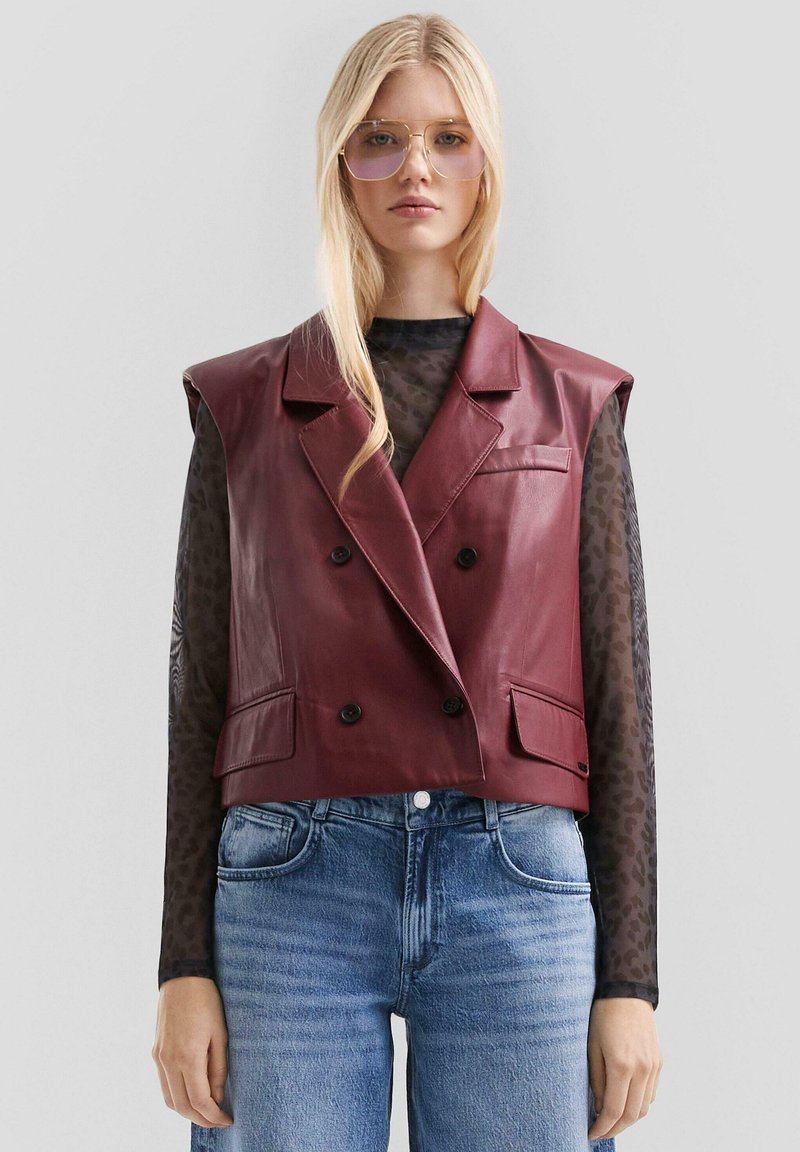 Bordeauxfarbenes Leder-Cropped-Blazer-Weste mit doppelt geknöpftem Design, spitzen Revers, zwei vorderen Taschen und einer glatten Textur. Kombiniert mit einem transparenten schwarzen Oberteil.