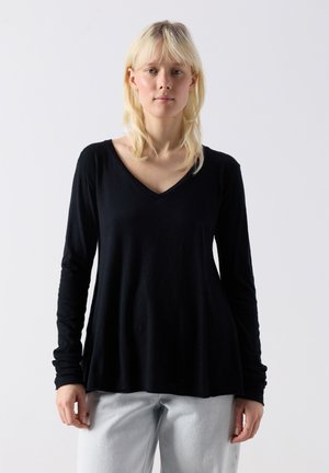 Lyshåret kvinde iført en løs sort langærmet V-hals top og lysegrå bukser, stående foran en ensartet lys baggrund.