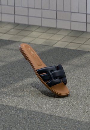 Braune flache Sandale mit schwarzen, ineinander verflochtenen Lederriemen auf strukturiertem grauem Pflasterboden und gefliestem Hintergrund.