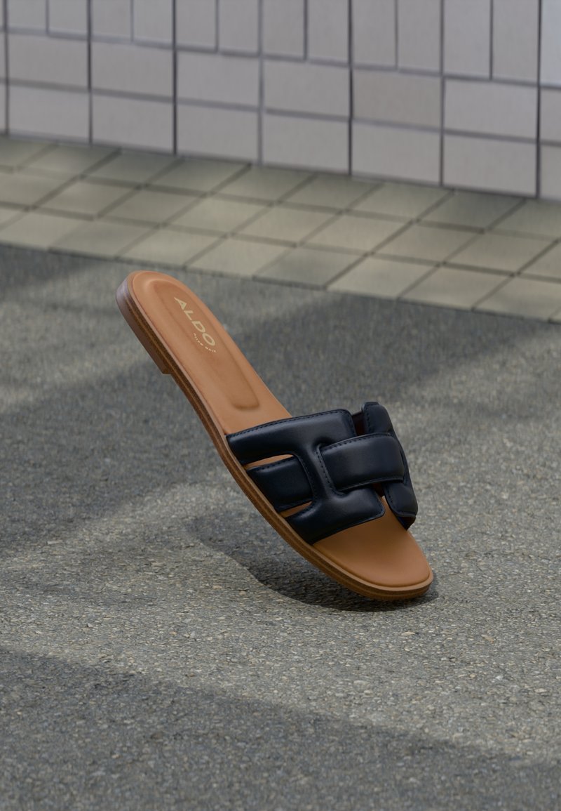 Sandalia plana marrón con tiras de cuero negras entrelazadas sobre pavimento gris texturizado y fondo de azulejos.