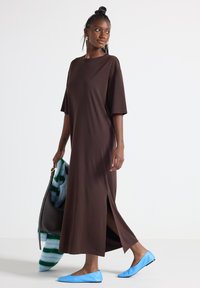 Brun, kortærmet maxi-kjole med sideslidser. Har blødt, glat stof. Bæres med lyseblå flade sko og holder en stribet blå-grøn tørklæde.