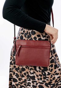 Bolso cruzado de piel roja con dos compartimentos con cremallera, correa ajustable y textura suave, combinado con una falda de estampado leopardo con pliegues.