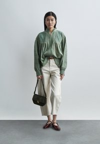 Γυναίκα φορώντας μπλούζα με κεντήματα σε χρώμα φασκόμηλο, κρεμ παντελόνι μέχρι τον αστράγαλο, μπορντό φλατ παπούτσια, κρατώντας μια σκουροπράσινη τσάντα, στέκεται μπροστά σε απλό φόντο.