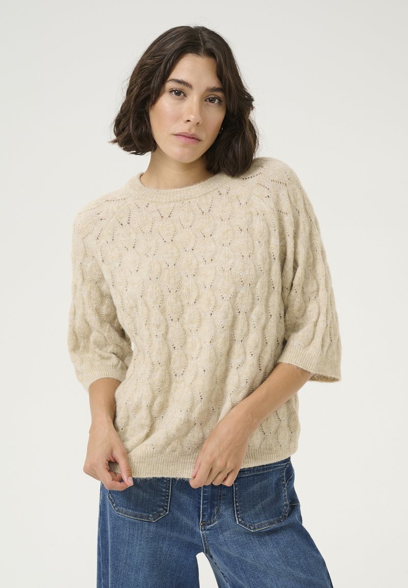 Beige gebreide trui met een kabelpatroon, ronde halslijn en korte mouwen. Gecombineerd met blauwe wijde jeans. Zachte textuur, ontspannen pasvorm.