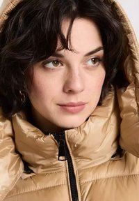 Brązowa kurtka puchowa z pikowanym wykończeniem i wysokim kołnierzem. Wyposażona w zamek i kaptur. Zbliżenie na osobę noszącą kurtkę.