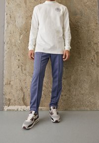 Long-sleeve white cotton shirt with a logo on the chest, paired with blue polyester pants and multicolor Nike sneakers.