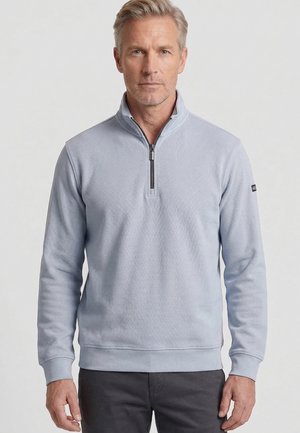 Man van middelbare leeftijd met licht haar, gekleed in een lichtgrijze kwart-rits trui en donkere broek, staand tegen een effen achtergrond.