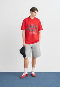 Νέος άνδρας με κόκκινο αθλητικό πουκάμισο Adidas, γκρι σορτς, κόκκινα αθλητικά παπούτσια Adidas, κρατώντας μαύρο καπέλο, στέκεται σε μπλε πάτωμα μπροστά από λευκό τοίχο.