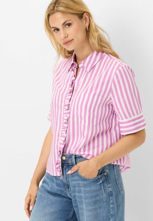 Femme aux cheveux blonds portant une chemise à manches courtes rayée verticalement rose et blanche et un jean bleu, une main dans la poche, légèrement de profil.