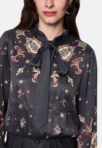 Schwarze, seidige Bluse mit Paisley- und Blumenmustern in Creme, Rot und Gold. Verfügt über eine Schleife am Hals und einen gerafften Taillendetail.