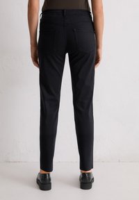 Persoon in zwarte rechte broek en zwarte instapschoenen die tegen een effen witte muur staat, gezien van achter.