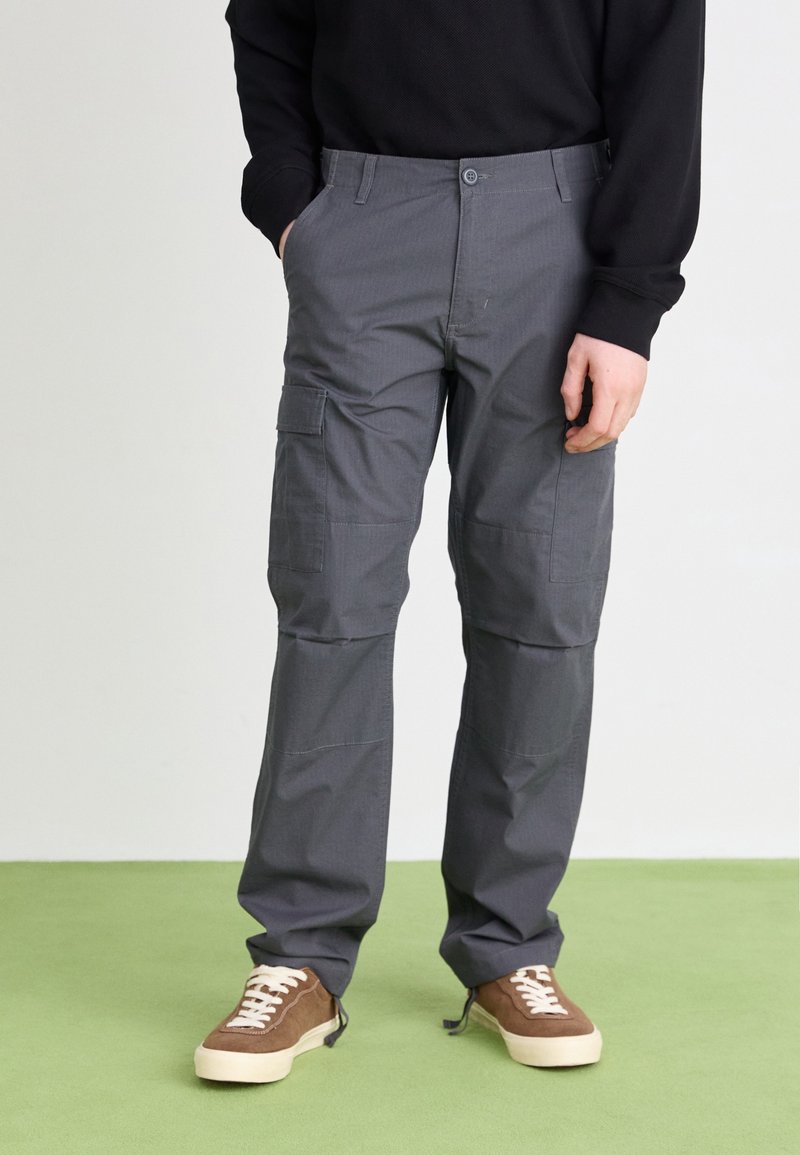 Persoon in grijze cargo broek en bruine sneakers met witte veters, staand op een groene vloer tegen een plain witte achtergrond.