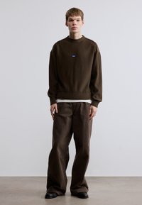 Pull en maille marron surdimensionné avec poignets et ourlet côtelés, arborant un petit patch logo bleu. Assorti à un pantalon marron ample et des chaussures noires.