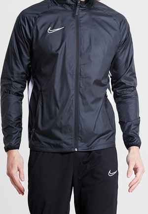 Mężczyzna ubrany w czarną kurtkę Nike na zamek i czarne spodnie Nike, stojący z ramionami swobodnie opuszczonymi wzdłuż ciała na jednolitym tle.