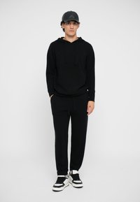 Hanorac cu glugă negru, cu buzunare frontale, purtat cu pantaloni joggers asortați și adidași sportivi negri și albi. Modelul stă în fața unui fundal alb.