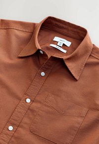 Chemise en coton slim fit marron avec un col boutonné, des boutons blancs et une poche poitrine. Les étiquettes indiquent la taille M et la marque NEXT.