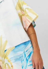 Camicia a maniche corte con una base bianca e un design colorato ad acquerello di foglie e onde in tonalità pastello di rosa, verde e blu.