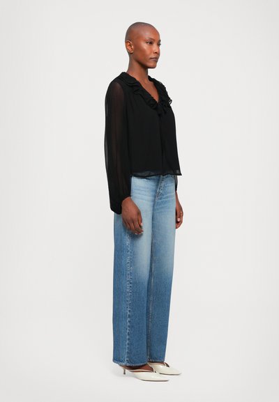 Zwarte sheer blouse met lange mouwen en ruches, gecombineerd met lichtblauwe high-waisted straight-leg jeans en witte puntige hakken.