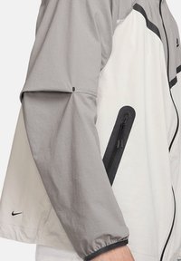 Lichte, twee-kleurige jas met een grijze bovenkant en een witte onderkant. Voorzien van een zwarte ritszak en elastische manchetten.