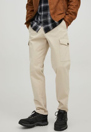 Pantalones cargo beige con bolsillos laterales, un ajuste relajado y una textura sutil, combinados con una camisa de cuadros en blanco y negro y botas negras.