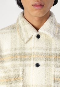 Fleece skjortejakke med et cremet og lysegrønt ternet mønster. Har krave, sorte knapper og en tekstureret, blød overflade.