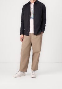 Camicia nera a maniche lunghe con bottoni sopra una maglietta grafica rosa chiaro, abbinata a pantaloni beige a gamba larga e sneaker bianche.