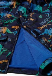 Granatowa kurtka z kolorowym wzorem dinozaurów w odcieniach pomarańczu, zieleni i niebieskiego. Wnętrze wyściełane miękką niebieską tkaniną, z zamkiem błyskawicznym.