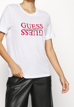 Person iført en hvid T-shirt med korte ærmer med rød "GUESS"-tekst spejlvendt og en vandret sølv blomsterdekoration, kombineret med sorte læderbukser.