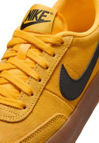 Sneaker gialla in suede con logo Nike nero, linguetta imbottita e lacci arancioni. La suola è in gomma traslucida, evidenziando i dettagli testurizzati.