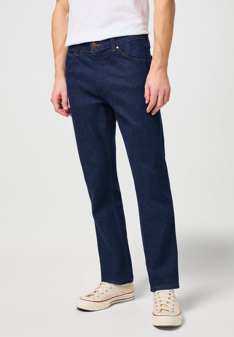 Donkerblauwe denim jeans met een rechte snit, vijf zakken en koperen knoopaccenten. Draagt met crèmekleurige sneakers.