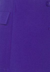 Lila tyg, slät textur, med en påsydd ficka och glänsande knappar. Rak silhuett med rena linjer och minimal detaljering.