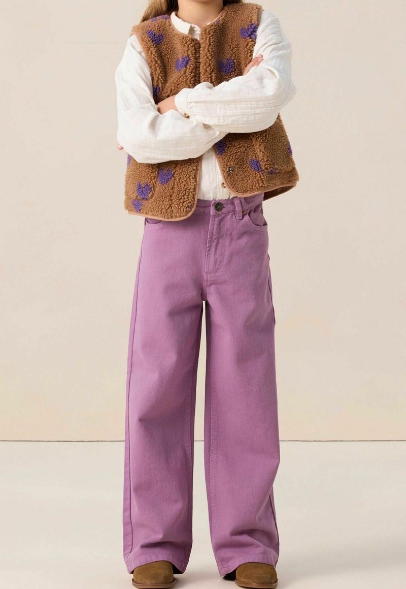 Enfant portant un gilet en polaire marron avec des cœurs violets, une chemise blanche à manches longues, un pantalon violet à jambes larges et des chaussures marron, bras croisés.
