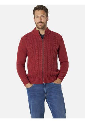 Mann trägt einen roten, kabelgestrickten Zip-Pullover mit Händen in den Taschen einer blauen Jeans, steht vor einem schlichten Hintergrund.