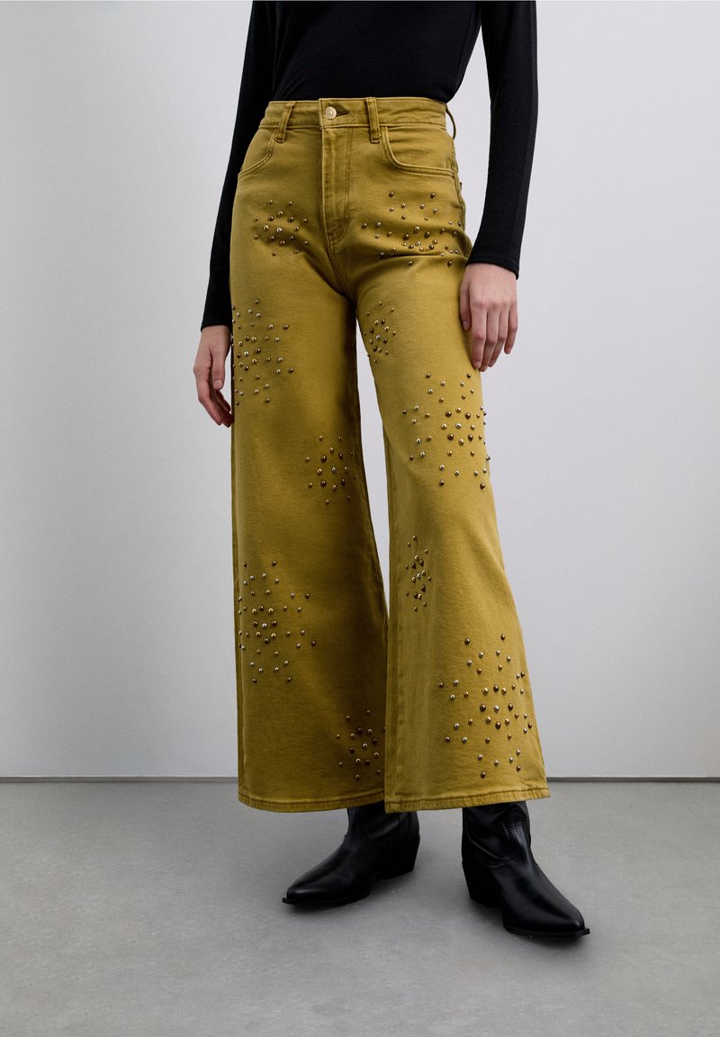 Pantalones de pierna ancha en algodón amarillo mostaza, con tachuelas metálicas en un patrón disperso. Combinados con botines negros.