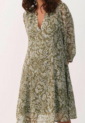 Vrouw draagt een olijfgroene jurk met wit bloemmotief, V-hals en driekwart doorzichtige mouwen, staand tegen een lichte achtergrond.