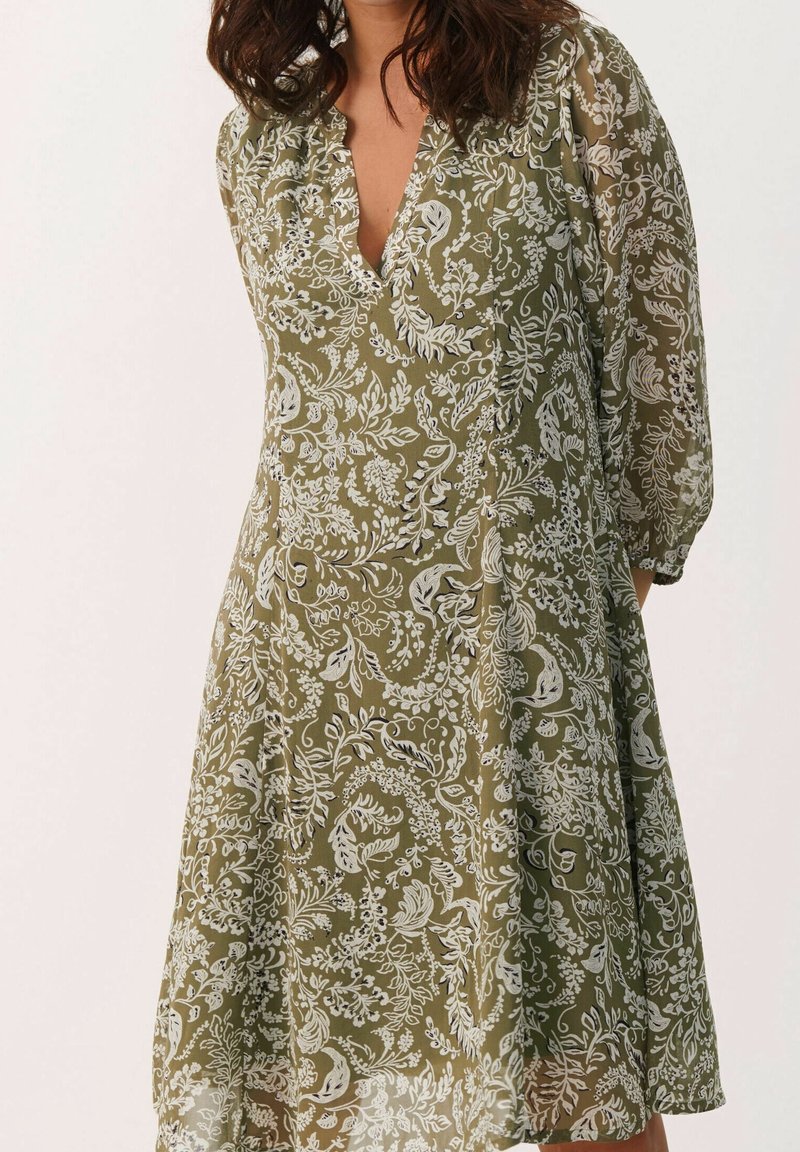 Femme portant une robe vert olive avec un motif floral blanc, col en V et manches trois-quarts transparentes, debout devant un fond clair.