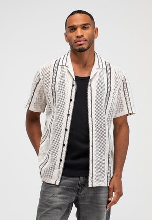 Man draagt een wit gebreid shirt met korte mouwen en zwarte verticale strepen over een zwart hemd, gecombineerd met grijze spijkerbroek, staat met één hand in zijn zak.