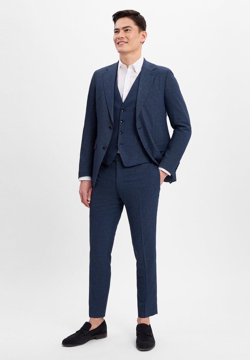 Dunkelblaues dreiteiliges Anzugset mit strukturierter Oberfläche, schmal geschnittene Hose und passender Weste. Schwarze Loafers runden das Outfit ab.
