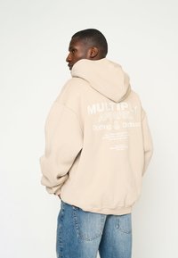 Hoodie oversize beige in tessuto texturizzato, con testo bianco stampato sul retro. Abbinato a jeans denim azzurri chiari.