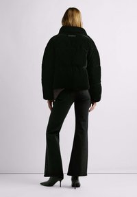 Melna pūkaina jaka ar augstu apkakli, ar kastveida siluetu un ķemmlaužu tekstūru, nēsāta kopā ar tumšām platām džinsa biksēm un potīšu zābakiem.