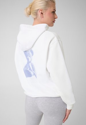 Femme blonde portant un sweat à capuche blanc avec un motif abstrait violet dans le dos et des leggings gris clair, debout devant un fond gris.