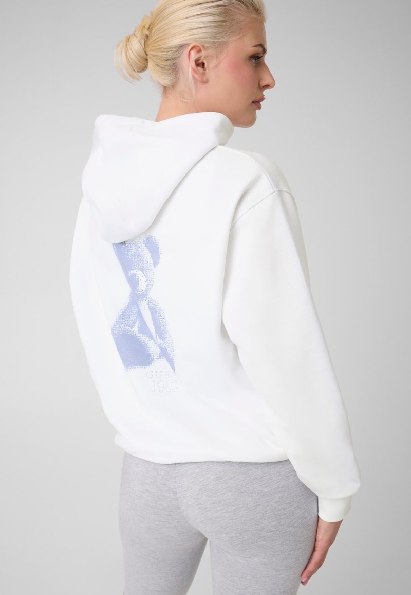 Femme blonde portant un sweat à capuche blanc avec un motif abstrait violet dans le dos et des leggings gris clair, debout devant un fond gris.